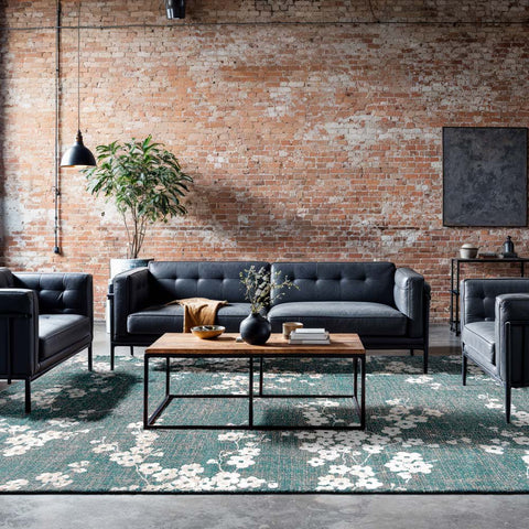 Tappeto floreale blu profondo con motivi petalo azzurri Lago Blu Sakura di Louis De Poortere posizionato al centro di un soggiorno moderno con divano grigio e tavolino in legno