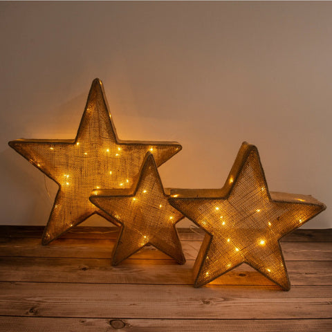 Set di tre stelle decorative a LED in tessuto e metallo marrone di varie dimensioni appoggiate sul pavimento in parquet di un soggiorno moderno in stile Japandi