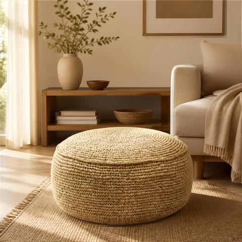 Pouf in rafia naturale Flores posizionato accanto a un divano chiaro in un soggiorno luminoso.