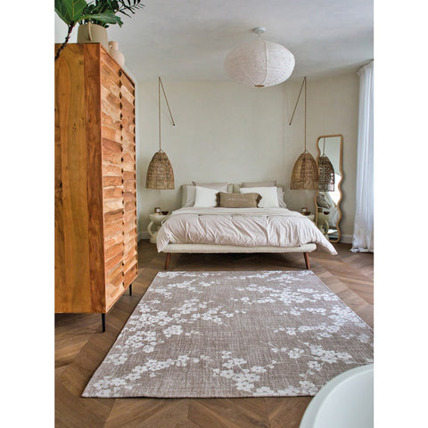 Tappeto flatweave ecologico con motivi floristici giapponesi sfumati nei toni del grigio e del marrone Sakura Morning Mist di Louis De Poortere steso su un pavimento in legno chiaro in camera da letto.