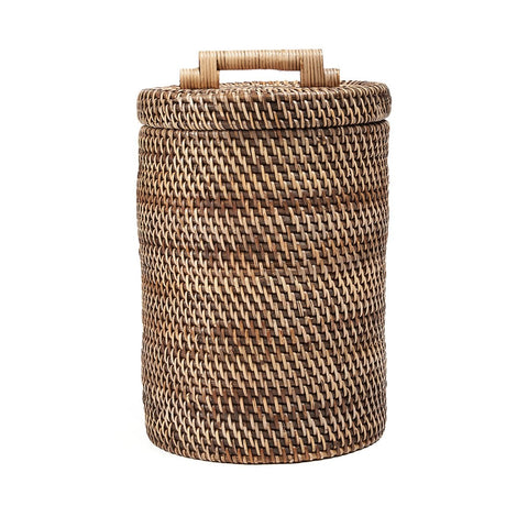 Cesto contenitore alto in rattan marrone naturale con coperchio posizionato accanto a una poltrona in un soggiorno luminoso
